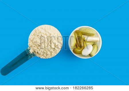 Protein powder in measuring plastic spoon and vitamin complex omega 3 glucosamine capsules supplements on blue background sports nutrition top view.
