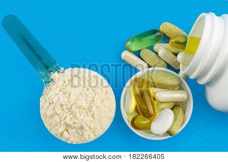 Protein powder in measuring plastic spoon and vitamin complex omega 3 glucosamine capsules supplements on blue background sports nutrition top view.