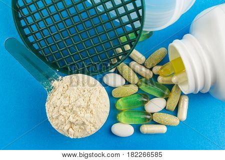 Protein powder in measuring plastic spoon and vitamin complex omega 3 glucosamine capsules supplements on blue background sports nutrition top view.