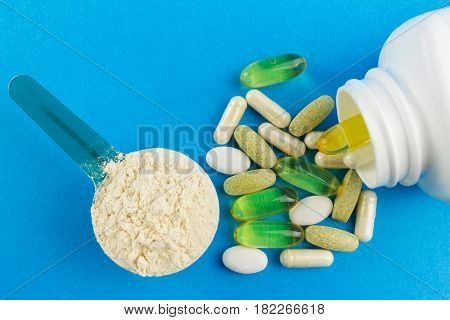 Protein powder in measuring plastic spoon and vitamin complex omega 3 glucosamine capsules supplements on blue background sports nutrition top view.
