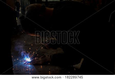 Skilled worksman welding wrought iron metal rods together in dark workshop with blue smoke and orange sparks wearing protective UV eyewear gear for eye protection