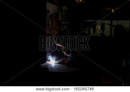 Worksman welding wrought iron metal rods together in dark workshop with blue smoke and orange sparks wearing protective UV eyewear gear for eye protection in very dark workshop