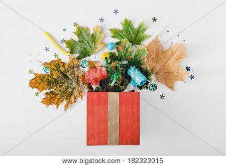Red gift box with various colorful dry leaves party confetti streamers noise makers and candles on a white background. Colorful celebration background. Flat lay