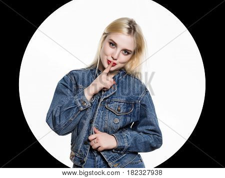 young blonde woman in denim jaket and jeans shows Index finger to the lips on white background. White circle on a black background