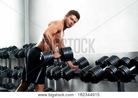 Handsome young shirtless man working out at the gym lifting heavy dumbbell looking to the camera confidently copyspace fitness workout muscles sport motivation effort achievement concept.