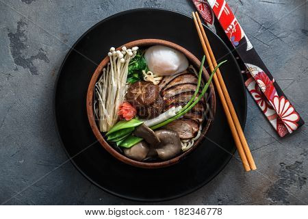 Ramen noodles with duck, egg, enoki and shiitake mushrooms with broth on dark background, top view
