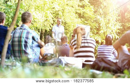 A group of young students in the park. View of a man gesticulating with his hands, standing against a defocused group of people sitting in front of him on the grass.