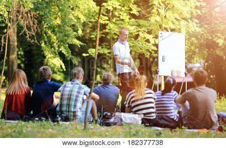 A group of young students in the park. View of a man gesticulating with his hands, standing against a defocused group of people sitting in front of him on the grass.