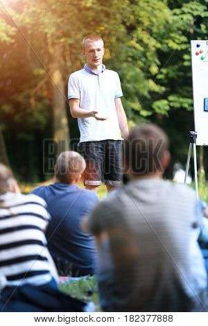 A group of young students in the park. View of a man gesticulating with his hands, standing against a defocused group of people sitting in front of him on the grass.
