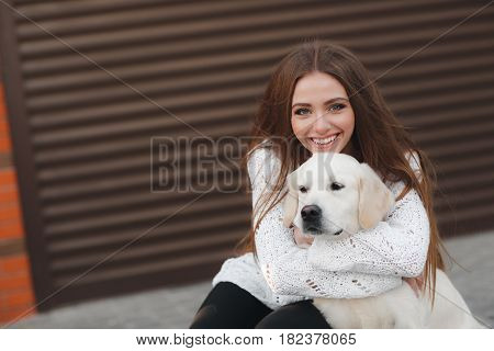 A young,beautiful woman with long,straight blond hair,a beautiful smile and white straight teeth,dressed in leggings black and white sweater posing outdoors in the spring,sitting in the city with his friend,an adult dog breed Golden Retriever