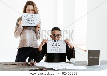 Woman holding sheet of paper and standing near the african man which sitting by the table and showing shet of paper