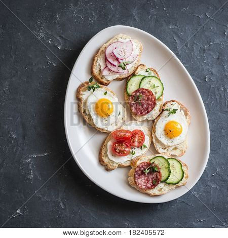 Variety of mini sandwiches with cream cheese vegetables quail eggs and salami. Sandwiches with cheese cucumber radish tomatoes salami quail eggs on a dark background top view
