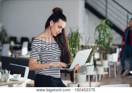 Attractive adult woman holding a laptop and typing a message to her subordinates to imform them about changes in schedule