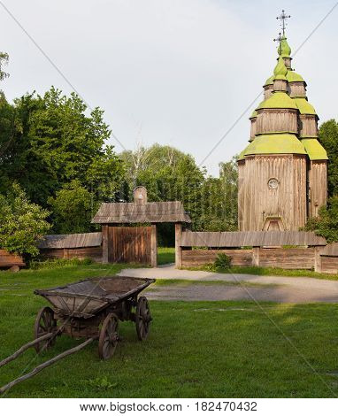 Ancient wooden church (XVIII century) from the Central Ukraine in the open-air Museum of Folk Architecture near Kyiv. Old wooden cart on the green grass in the foreground.