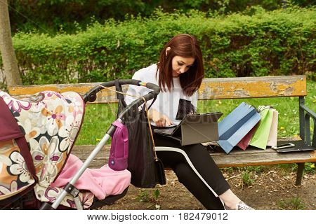 A woman with baby carriage and shopping bags working with tablett in the park while sitting on a bench.