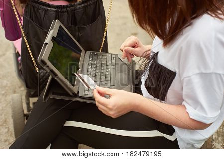 Crop shot of a woman with baby carriage holding in her hand a credit card and making online shopping with tablett in the park while sitting on a bench.
