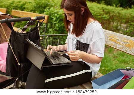 A woman with baby carriage holding in her hand a credit card and making online shopping with tablett in the park while sitting on a bench.