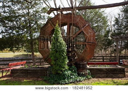 A large unused wooden paddlewheel surrounded by flower beds starting to bud and an evergreen tree in a spring landscape