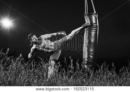 Black and white shot of a professional kick boxer practicing on a heavy punching bag outdoors at night copyspace motivation lifestyle workout muscles power strength masculinity martial sportsperson.