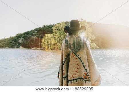 Boho style woman wearing in poncho and hat walking on lake rear view.