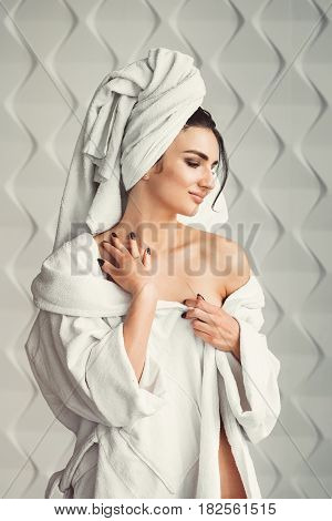 sexy young girl with dark hair, big eyes and dark eyebrows wearing white bath robe whith towel on her head. Model with light nude make-up, white studio background, beauty photo