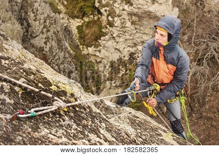 The Young Man climbs the rock on a rope with safety belts, insurance and rope, in full mountaineering equipment and a helmet and hood