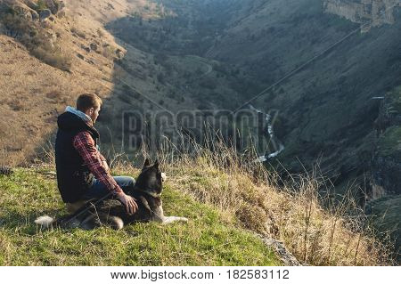 A man with a beard walking his dog in the nature, standing with a backlight at the rising sun, casting a warm glow and long shadows against the background of the gorge and trees on caucazus