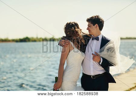 Groom Holds Bride's Veil Blown By The Wind Hugging Her Tender