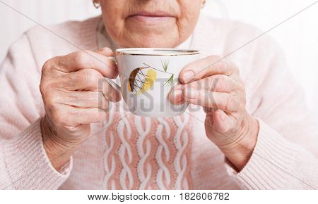 An elderly woman drinks tea at home. Senior woman holding cup of tea in their hands at table close-up