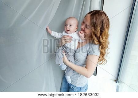 Young mother holding her newborn child. Woman and new born boy relax in a white bedroom near the windiow. Mother breast feeding baby. Family at home. Young mother playing whith her newborn son