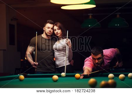 Group of young adults having fun in club playing billiard game. Young man aiming to take a snooker shot while young romantic couple hugging.