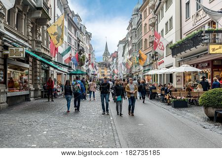ZURICH SWITZERLAND - MAY 14 2016: People on shopping street Rennweg with boutique shops flags on old buildings and St. Peter church clock tower in Zurich Switzerland.