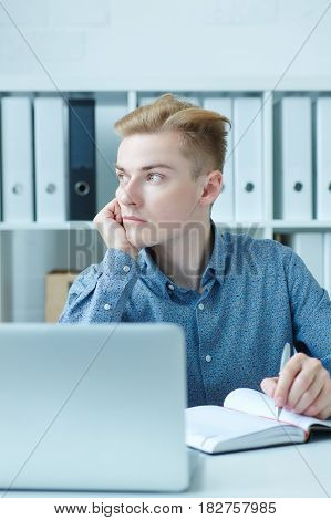 Handsome pensive young business man sitting at the dask with laptop and looking at the window.