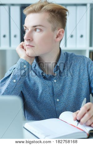 Handsome pensive young business man sitting at the dask with laptop and looking at the window.