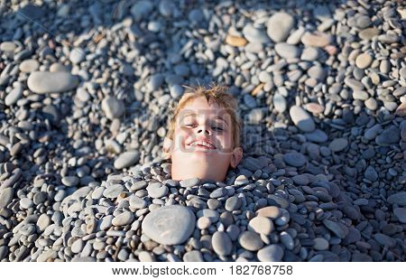 A young man with light red hair laughs. His body was buried under stones on the beach only his head sticking out the sun is shining