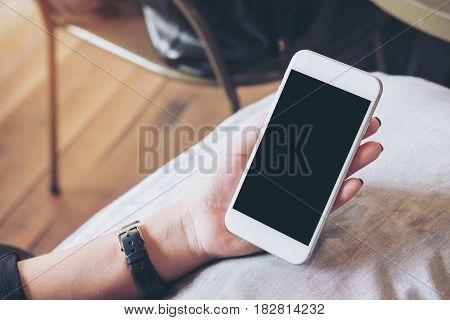Mockup image of hands holding white mobile phone with blank black screen on pillow with wooden floor background