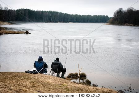 CARNIKAVA LATVIA - APRIL 1 2017: Two unknown men with a pole catch fish from the river shore.