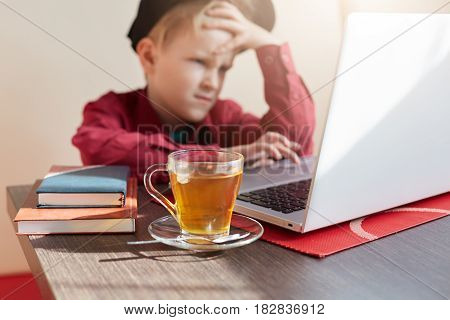 An Emotional Llitle Boy Wearing Trendy Clothes And Cap Sittiing At The Table Working With His Laptop