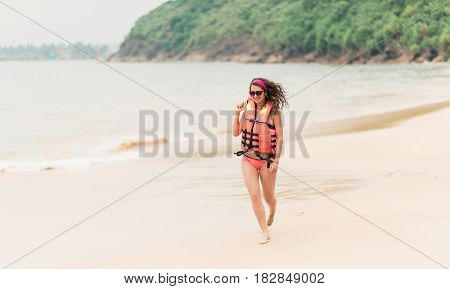 A girl in a life jacket and glasses is running along a deserted beach. A beautiful curly girl with a bright manicure in an orange life jacket monitors the safety on the beach.