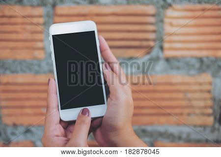 Mockup image of hand holding white mobile phone with blank black screen and brick wall background
