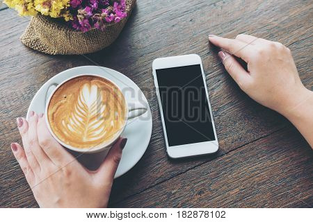 Mockup image of white mobile phone with blank black screen and hand holding hot latte coffee on vintage wood table in cafe