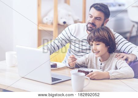 As like as two peas. Riant boy and his dad wearing casual clothes and watching football match at laptop while spending free time in the bosom of their family