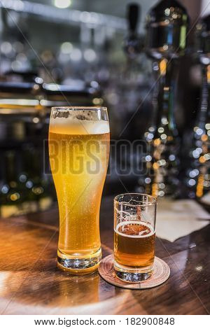 Light beer in a beer glass and dark beer in a small glass lighted with warm light on a wooden bar counter. Vertical frame