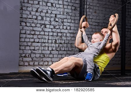 Happy father and little son exercising with gimnastic rings and smiling against brick wall in the gym. Fathet and son in the sportwear and sportive shoes. Happy family. Rearing child in the healthy lifestyle.