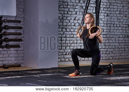Young fit woman in the sportwear doing pull-ups on gymnastic rings against brick wall in the cross fit gym. Sporty woman in the black t-shirt, leggings and sportive shoes. Portrait of beautiful sporty woman. Healthy lifestyle.