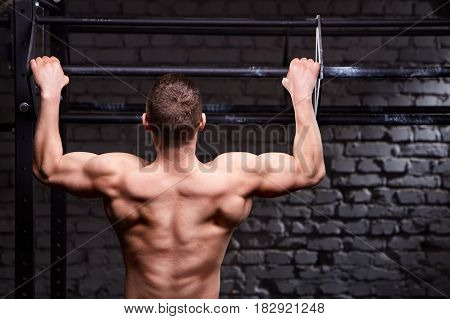 Rear view photo of muscular male doing exercises on horizontal bar against brick wall at the cross fit gym. Body and muscle. Power and energy. Healthy lifestyle.