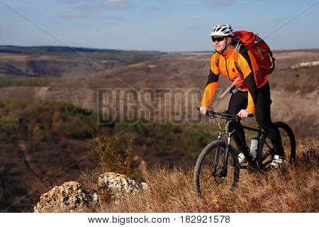 Cyclist in the orange jacket standing with his bike on the top of a hill against beautiful landscape. Sky, hill and rock in the countryside. Sportsman in the helemet, sunglasses and with red backpack. Extreme travel in the summer season.