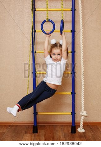 Little girl in sports clothes doing exercises on sports rings on a Swedish wall. The concept of a healthy lifestyle from a young age. Children sport.