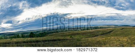 A dirt road through a meadow, under a mighty sky, near the mountains.