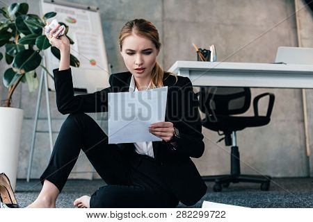 Barefoot Businesswoman Sitting On Floor Near Work-table With Crumpled Paper And Reading Documents